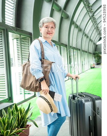 Asian mature woman in her 60s smiling and standing with suitcase duffel bag and holding boarding pass and passport for travel after covid19 94528998