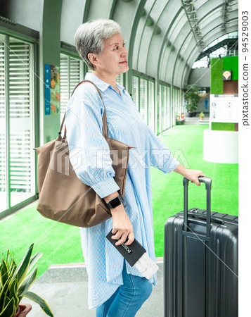 60多歲的亞洲熟年婦女微笑著站著,身旁有手提箱行李袋和手持登機證與護照,適用於covid19疫情後旅 60多歲的亞洲熟年婦女微笑著站著,身旁有手提箱行李袋和手持登機證與護照,適用於covid19疫情後旅 94529003