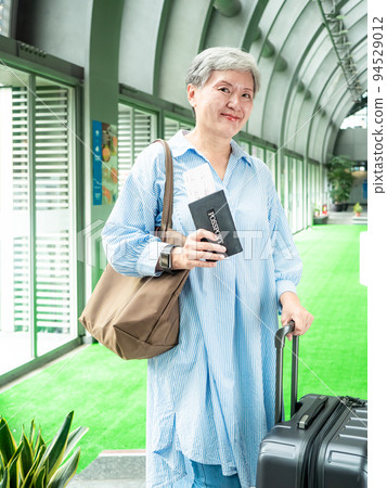 60多歲的亞洲熟年婦女微笑著站著，身旁有手提箱行李袋和手持登機證與護照，適用於covid19疫情後旅 94529012
