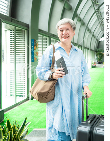 60多歲的亞洲熟年婦女微笑著站著，身旁有手提箱行李袋和手持登機證與護照，適用於covid19疫情後旅 94529013