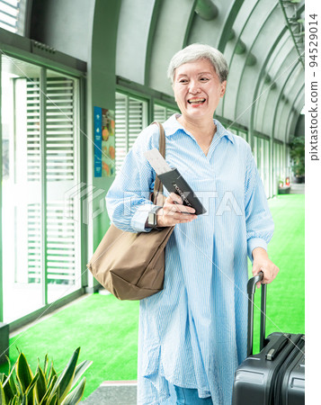 60多歲的亞洲熟年婦女微笑著站著，身旁有手提箱行李袋和手持登機證與護照，適用於covid19疫情後旅 94529014