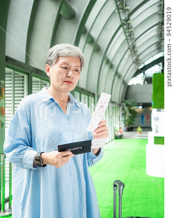 60多歲的亞洲熟年婦女微笑著站著，身旁有手提箱行李袋和手持登機證與護照，適用於covid19疫情後旅 94529019