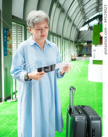 60多歲的亞洲熟年婦女微笑著站著，身旁有手提箱行李袋和手持登機證與護照，適用於covid19疫情後旅 94529023