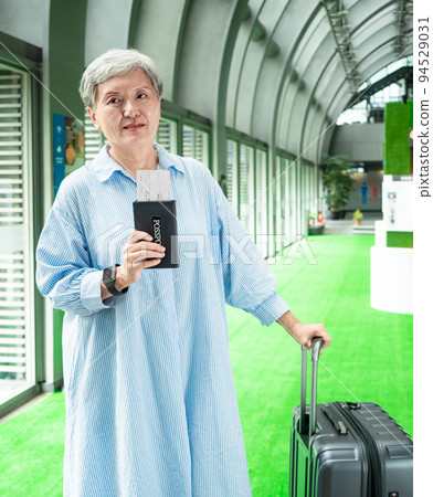 60多歲的亞洲熟年婦女微笑著站著，身旁有手提箱行李袋和手持登機證與護照，適用於covid19疫情後旅 94529031