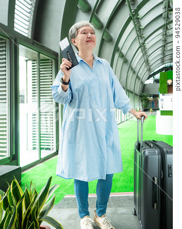 60多歲的亞洲熟年婦女微笑著站著，身旁有手提箱行李袋和手持登機證與護照，適用於covid19疫情後旅 94529038