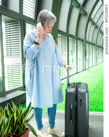 60多歲的亞洲熟年婦女微笑著站在身旁有手提箱行李袋和使用智能手機，適用於covid19疫情後旅行的概 94529063