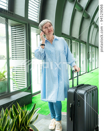 60多歲的亞洲熟年婦女微笑著站在身旁有手提箱行李袋和使用智能手機，適用於covid19疫情後旅行的概 94529066