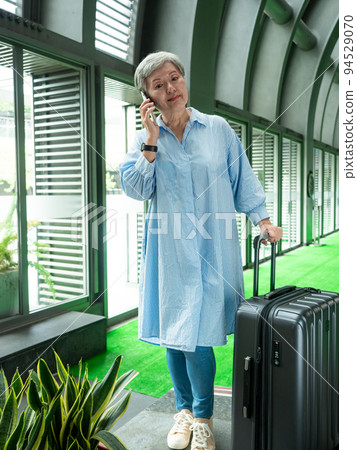 60多歲的亞洲熟年婦女微笑著站在身旁有手提箱行李袋和使用智能手機，適用於covid19疫情後旅行的概 94529070