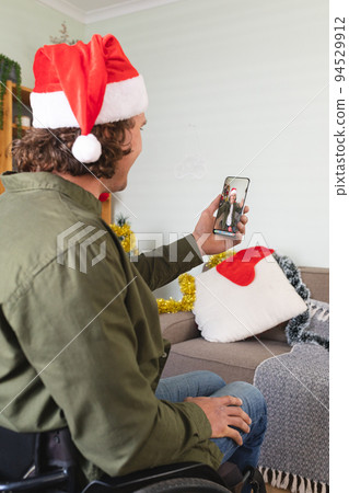 Caucasian man in wheelchair and santa hat making christmas smartphone video call with male friend Caucasian man in wheelchair and santa hat making christmas smartphone video call with male friend 94529912