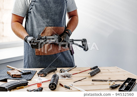 Close up of young man in apron disassembling a gun above the table 94532560