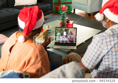 African american senior couple in santa hats on christmas laptop video call with friend 94534328
