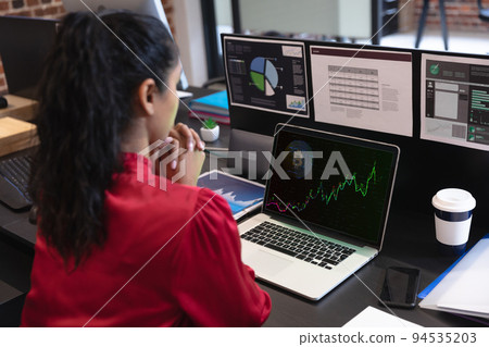 Biracial businesswoman sitting at desk using laptop with graph and statistical data on screen 94535203