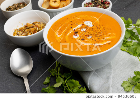 Horizontal image of bowl of tomato soup, with toast, seeds, leaves and peppercorns on slate 94536605