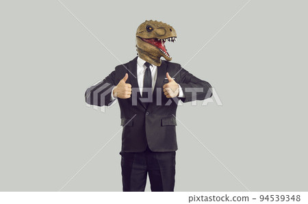 Funny businessman in humorous trendy dinosaur mask showing thumbs up on gray background. 94539348