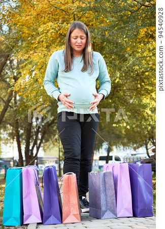 Unhappy pregnant woman with paper bags feeling stress during premature childbirth in city park. Young mother holding belly abdomen worried about pain after Black Friday shopping. Unhappy pregnant woman with paper bags feeling stress during premature childbirth in city park. Young mother holding belly abdomen worried about pain after Black Friday shopping. 94540928