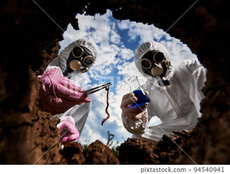 View from inside pit of researchers in protective overalls and gas masks studying soil and its inhabitants. One laboratory assistant holding tweezers with soil worm, the other glass flask with liquid. View from inside pit of researchers in protective overalls and gas masks studying soil and its inhabitants. One laboratory assistant holding tweezers with soil worm, the other glass flask with liquid. 94540941