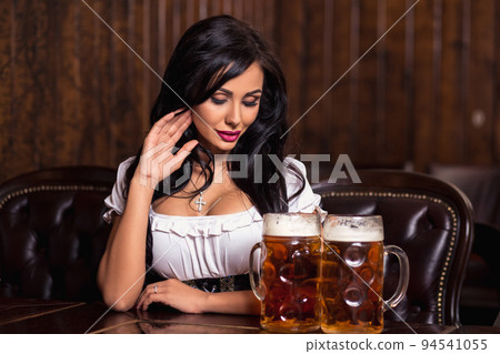 Oktoberfest woman wearing a traditional Bavarian dress dirndl posing with a beer mugs at bar 94541055