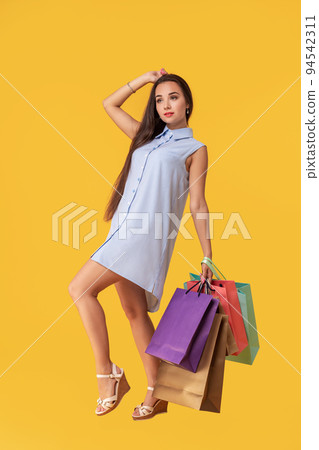 Full length photo of young woman in dress looking back on copy space and holding colorful shopping packages over yellow background 94542311