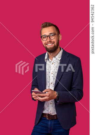Young cheerful lawyer or businessman is standing on the pink background and smiling, reading good news on internet at his mobile phone. Young cheerful lawyer or businessman is standing on the pink background and smiling, reading good news on internet at his mobile phone. 94542362