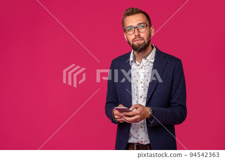 Young cheerful lawyer or businessman is standing on the pink background and smiling, reading good news on internet at his mobile phone. Young cheerful lawyer or businessman is standing on the pink background and smiling, reading good news on internet at his mobile phone. 94542363