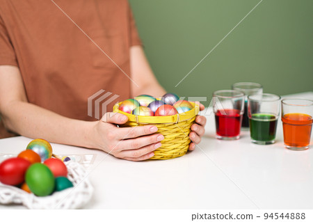 A young beautiful woman in a brown t-shirt paints Easter eggs in different colors. Preparing Easter Eve at home. Green wall. 94544888