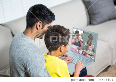 Biracial boy with father using laptop for video call, with elementary school pupils on screen 94545195