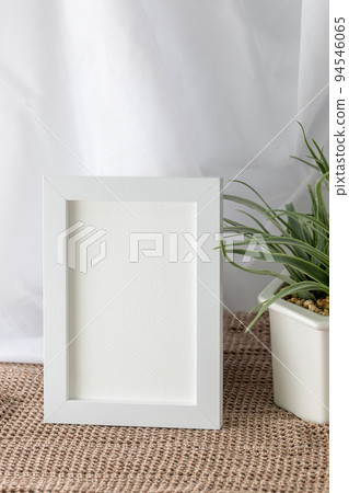 Blank vertical white frame beside a houseplant, painting or artwork display on brown knitted cloth 94546065