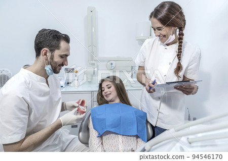 Young woman is sitting in dental chair in clinic, office. Man doctor is showing jaw Young woman is sitting in dental chair in clinic, office. Man doctor is showing jaw 94546170