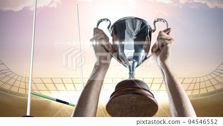 Composition of male rugby player celebrating victory, holding trophy on rugby pitch at stadium 94546552