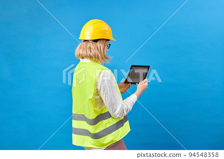 Woman repairman holding tablet. Girl engineer on studio blue background. Repairman uses tablet computer while working. Woman repairman looks up on the graph jn tablet. Foreman career Woman repairman holding tablet. Girl engineer on studio blue background. Repairman uses tablet computer while working. Woman repairman looks up on the graph jn tablet. Foreman career 94548358