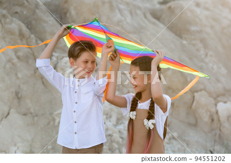 A girl and a boy carry a bright colored kite over their heads while walking in the mountains 94551202
