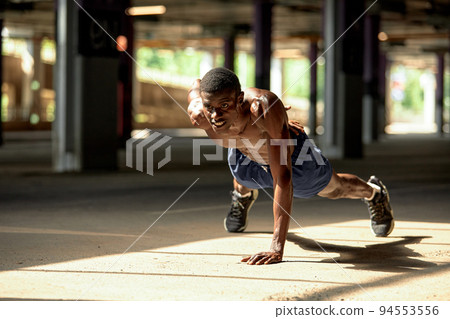 Side view of muscular black man in sportswear doing push ups during workout on street of modern downtown with urban background Side view of muscular black man in sportswear doing push ups during workout on street of modern downtown with urban background 94553556