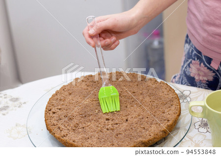 Working process of baking cake at home. Close up of female hand with silicone brush preparing chocolate biscuit cake. Culinary and delicious dessert concept. Working process of baking cake at home. Close up of female hand with silicone brush preparing chocolate biscuit cake. Culinary and delicious dessert concept. 94553848