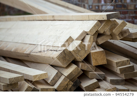 Close-up of piled stack of natural brown uneven rough wooden boards lit by bright sun. Industrial timber for carpentry, building, repairing and furniture, lumber material for construction. Close-up of piled stack of natural brown uneven rough wooden boards lit by bright sun. Industrial timber for carpentry, building, repairing and furniture, lumber material for construction. 94554279