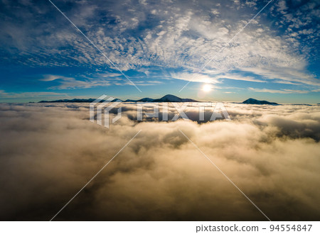 Aerial view of vibrant sunrise over white dense clouds with distant dark mountains on horizon. 94554847