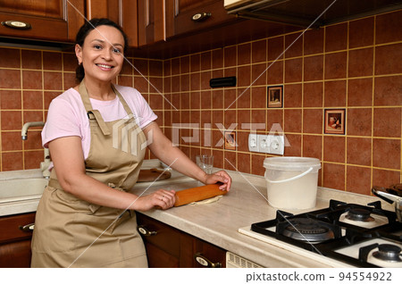 African American pretty woman, a housewife, chef confectioner wearing a beige chef's apron, rolling out the dough in the home kitchen, smiles cutely a beautiful toothy smile, looking at the camera African American pretty woman, a housewife, chef confectioner wearing a beige chef's apron, rolling out the dough in the home kitchen, smiles cutely a beautiful toothy smile, looking at the camera 94554922