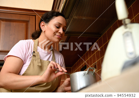 Close-up of a dark-haired beautiful middle-aged woman, a pleasant housewife, mixing ingredients in a saucepan, standing by the stove in home kitchen, preparing caramelized cherries for a homemade pie 94554932