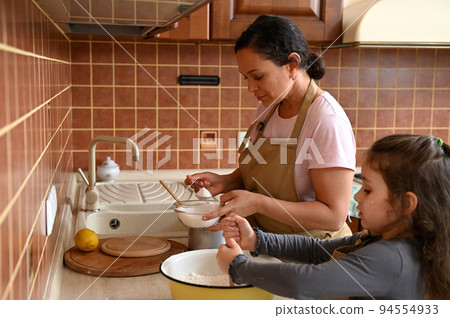 Happy family. Beautiful stunning dark-haired Hispanic woman housewife, loving mom and daughter, both in beige chef apron, preparing dough in the kitchen, enjoying cooking homemade festive pie together 94554933