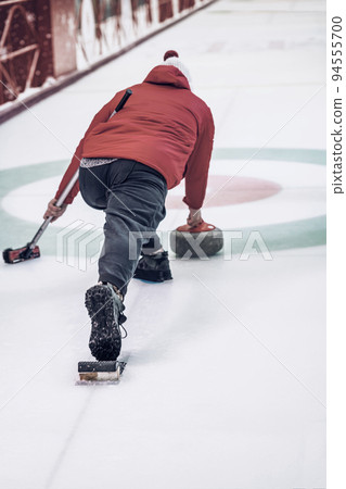 Curling game. Rear view of player playing curling on ice rink with granite stone and broom 94555700