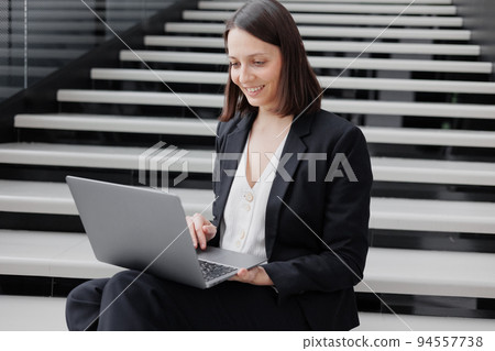 business woman in a formal suit with a laptop in an office or business center sits on the steps in the lobby and works online 94557738