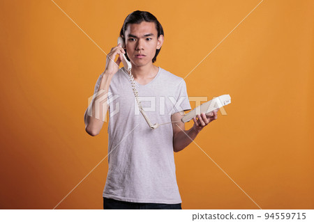 Young scared man talking on landline phone. Confused asian teen chatting, holding retro telephone, frightened person with terrified facial expression answering call on orange background 94559715