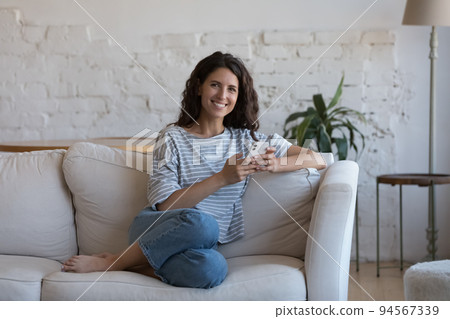Hispanic woman rest on sofa holds smartphone look at camera Hispanic woman rest on sofa holds smartphone look at camera 94567339