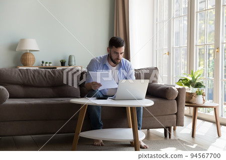 Serious focused millennial homeowner man checking paper bills Serious focused millennial homeowner man checking paper bills 94567700