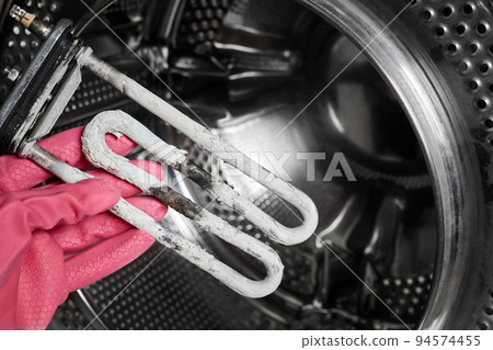 A man holds in his hand a burnt-out heating element of a washing machine against the background of a washing machine drum A man holds in his hand a burnt-out heating element of a washing machine against the background of a washing machine drum 94574455