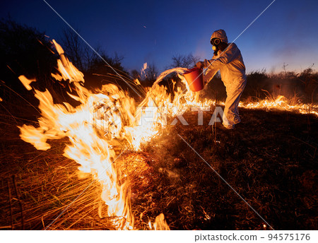 Fireman extinguishing fire in field with blue night sky on background. Man in protective radiation suit and gas mask pouring water on burning grass with smoke. Natural disaster concept. Fireman extinguishing fire in field with blue night sky on background. Man in protective radiation suit and gas mask pouring water on burning grass with smoke. Natural disaster concept. 94575176