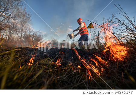 Firefighter ecologist extinguishing fire in field with evening sky on background. Male environmentalist holding bucket and pouring water on burning dry grass near warning sign with exclamation mark. Firefighter ecologist extinguishing fire in field with evening sky on background. Male environmentalist holding bucket and pouring water on burning dry grass near warning sign with exclamation mark. 94575177