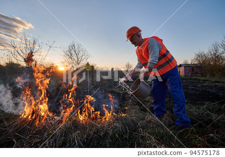 Fireman ecologist extinguishing fire in field with warning sign with exclamation mark end evening sky on background. Male environmentalist holding bucket and pouring water on burning dry grass. Fireman ecologist extinguishing fire in field with warning sign with exclamation mark end evening sky on background. Male environmentalist holding bucket and pouring water on burning dry grass. 94575178