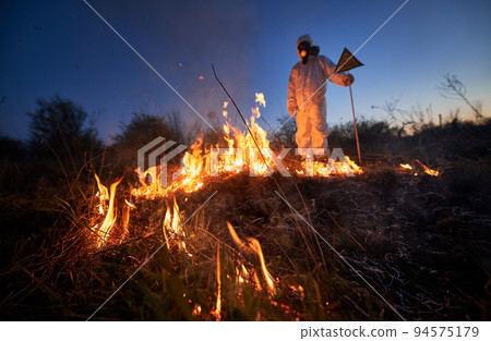 Fireman ecologist working in field with wildfire at night. Man in protective radiation suit and gas mask near burning grass with smoke, holding warning sign. Natural disaster concept. Fireman ecologist working in field with wildfire at night. Man in protective radiation suit and gas mask near burning grass with smoke, holding warning sign. Natural disaster concept. 94575179