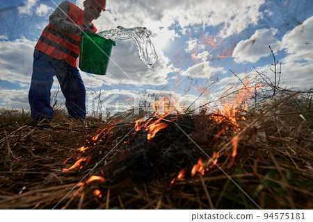 Firefighter ecologist fighting fire in field with cloudy sky on background. Cropped view of male environmentalist holding bucket and pouring water on burning dry grass. Natural disaster concept. Firefighter ecologist fighting fire in field with cloudy sky on background. Cropped view of male environmentalist holding bucket and pouring water on burning dry grass. Natural disaster concept. 94575181