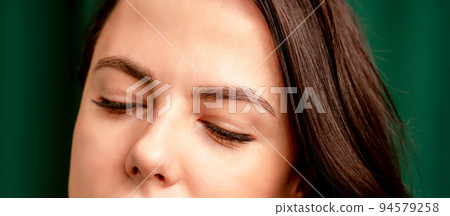 The fashionable young woman. Portrait of the beautiful female model with long hair and makeup with closed eyes. Beauty young woman with a brown curly hairstyle on the background of a green curtain The fashionable young woman. Portrait of the beautiful female model with long hair and makeup with closed eyes. Beauty young woman with a brown curly hairstyle on the background of a green curtain 94579258